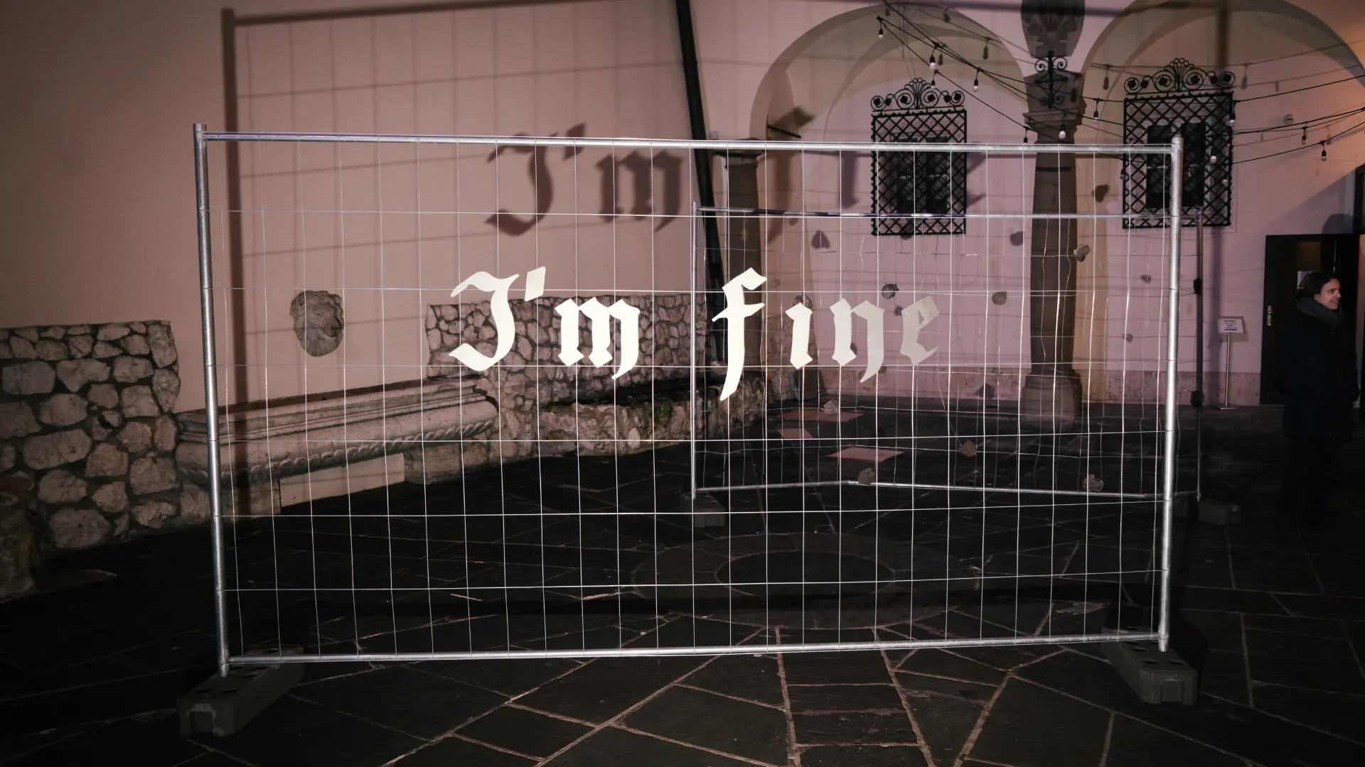 Metal gate featuring the repeated words I'm Fine displayed against a brick wall in a gallery.