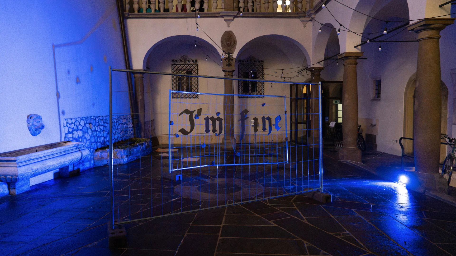Metal gate with the words I'm Fine displayed under blue lighting in an arched historic courtyard.