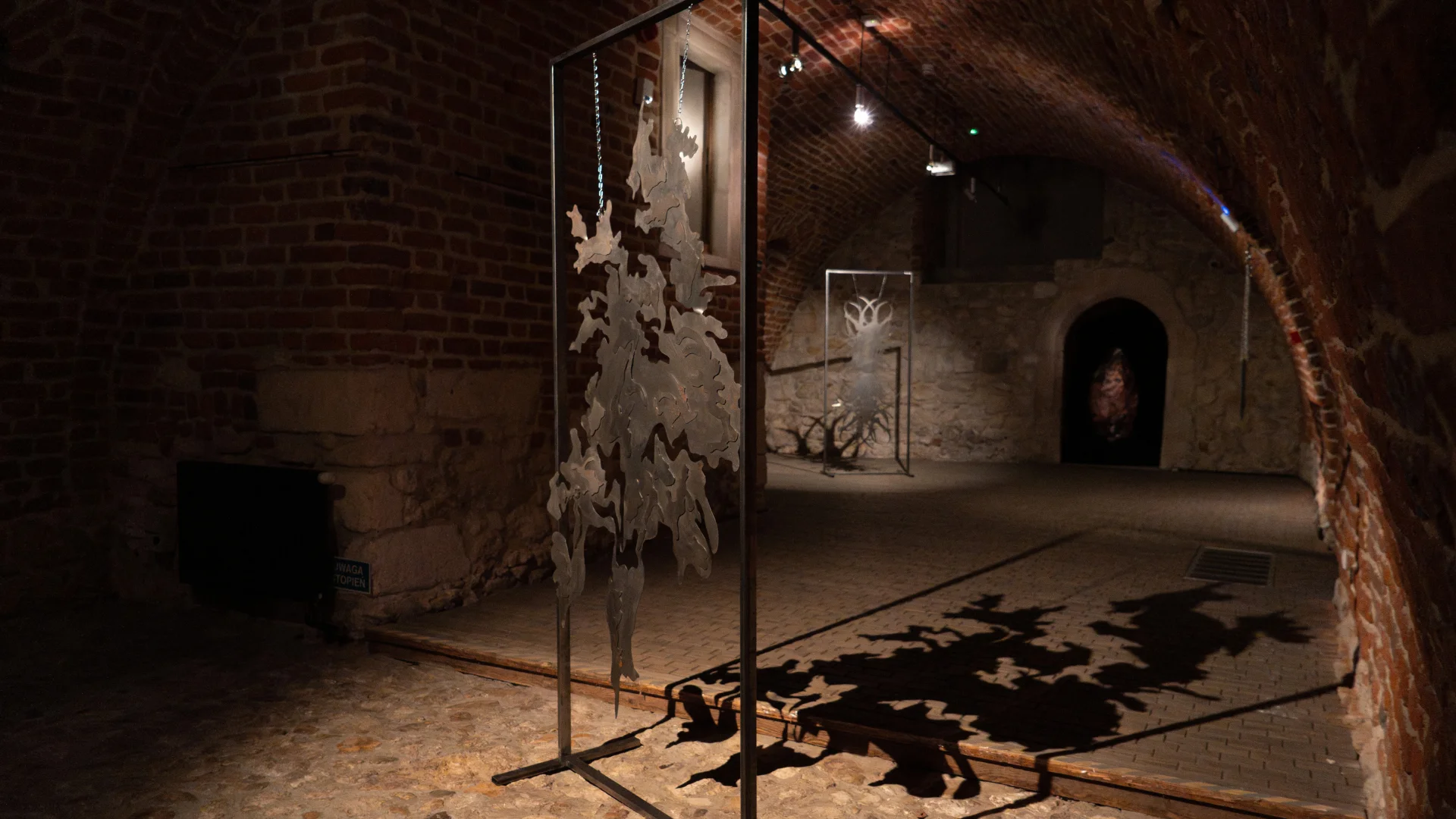 Suspended metal sculptures displayed in a brick-vaulted gallery corridor with dramatic lighting.