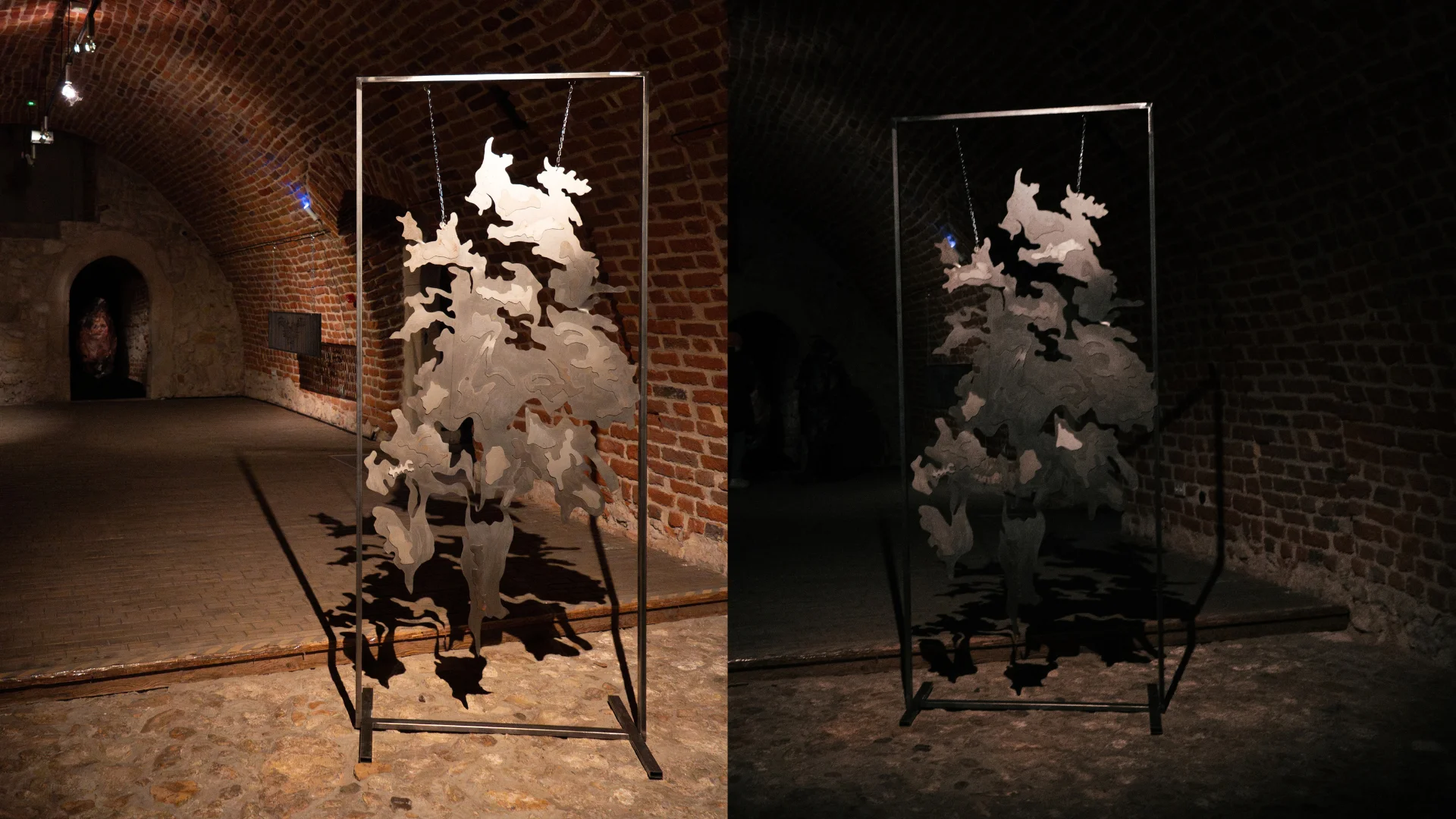 Two suspended abstract metal sculptures displayed inside rectangular frames in a dark brick gallery.