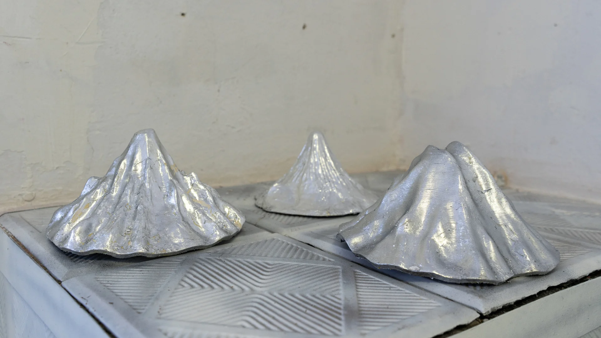 Three silver mountain-like sculptural objects with ridged surfaces arranged on a pale display platform.